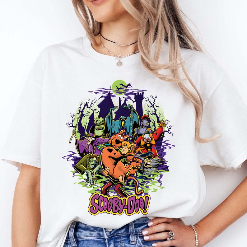 May include: A white t-shirt featuring a colorful graphic print of Scooby-Doo characters and various monsters in front of a dark castle silhouette. The design includes a green moon, purple smoke, and the text Scooby-Doo in a stylized yellow font.
