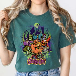May include: A dark green t-shirt featuring a graphic print of Scooby-Doo and Shaggy Rogers surrounded by various cartoon monsters, ghosts, and a haunted castle silhouette. The text Scooby-Doo is printed in yellow at the bottom of the graphic.