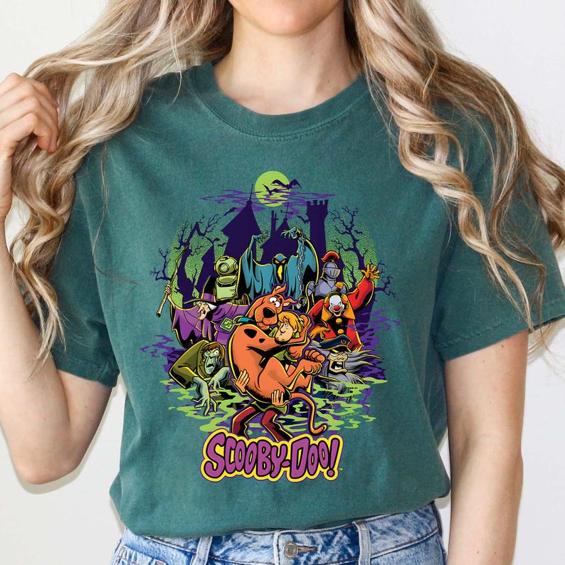 May include: A dark green t-shirt featuring a graphic print of Scooby-Doo and Shaggy Rogers surrounded by various cartoon monsters, ghosts, and a haunted castle silhouette. The text Scooby-Doo is printed in yellow at the bottom of the graphic.