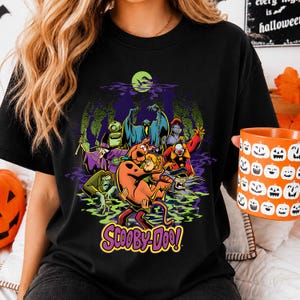 May include: A black t-shirt featuring a graphic print of Scooby-Doo and Shaggy surrounded by various classic villains from the series. The design includes a green moon, purple smoke, and the Scooby-Doo logo in yellow and purple lettering at the bottom.