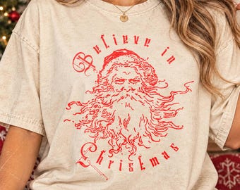 May include: A beige short-sleeve t-shirt featuring a red graphic of Santa Claus with a long beard. The text "Believe in Christmas" is printed in red lettering around the illustration. The fabric has a mottled, vintage-style texture.