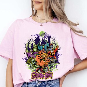 May include: A light pink t-shirt featuring a graphic print of Scooby-Doo and Shaggy surrounded by various cartoon monsters and a haunted castle silhouette. The Scooby-Doo logo is printed in yellow and purple at the bottom of the graphic design.