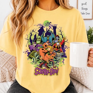 May include: A pale yellow t-shirt featuring a colorful graphic of Scooby-Doo and Shaggy surrounded by various ghosts and monsters in front of a dark castle silhouette. The text Scooby-Doo is printed in purple and yellow at the bottom of the design.