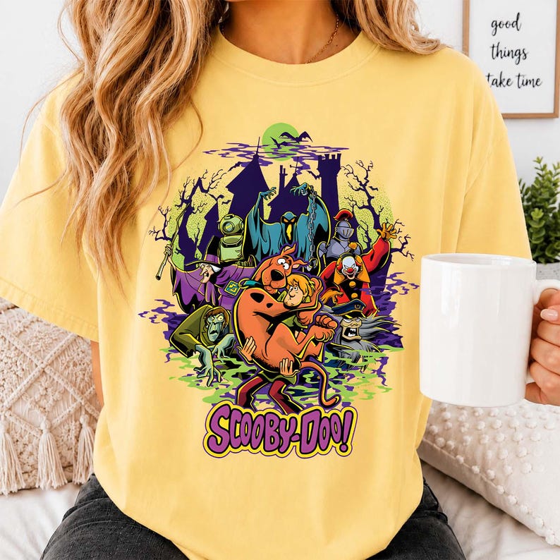 May include: A pale yellow t-shirt featuring a colorful graphic of Scooby-Doo and Shaggy surrounded by various ghosts and monsters in front of a dark castle silhouette. The text Scooby-Doo is printed in purple and yellow at the bottom of the design.