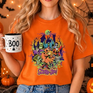 May include: An orange t-shirt featuring a graphic print of Scooby-Doo and various villains in front of a haunted castle under a green moon. The shirt is paired with a white ceramic mug that has the word BOO and small black stars printed on it.