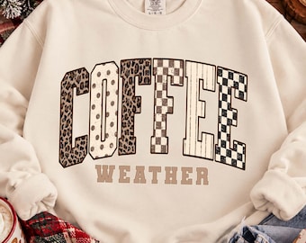 May include: A cream-colored crewneck sweatshirt featuring the word COFFEE in large, block-style letters with varied patterns including leopard print, polka dots, and checkerboard. Below, the word WEATHER is printed in a simple brown font.