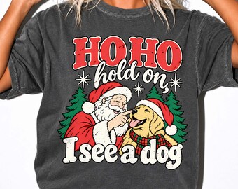 May include: A charcoal gray t-shirt featuring a graphic of Santa Claus touching the nose of a golden retriever wearing a red hat and plaid scarf. The text reads HO HO hold on, I see a dog, surrounded by green pine trees and white stars.