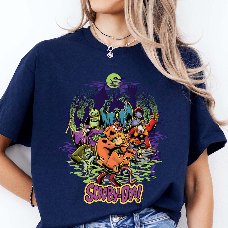 May include: A navy blue short-sleeve t-shirt featuring a colorful graphic print of Scooby-Doo and Shaggy surrounded by various cartoon villains in a swampy setting under a green moon. The text Scooby-Doo is printed in yellow at the bottom.