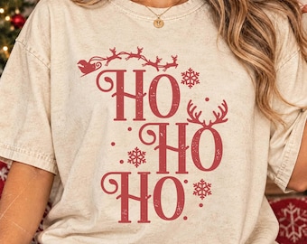 May include: A beige short-sleeved t-shirt featuring a red graphic print with the text HO HO HO. The design includes a silhouette of Santa Claus in a sleigh pulled by reindeer, snowflake icons, and a pair of reindeer antlers on the letter O.