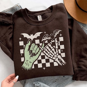 May include: A dark brown crewneck sweatshirt featuring a graphic print of a light green hand and a white skeleton hand performing a pinky promise. The design includes a black and white checkered background, two flying bats, and small stars.