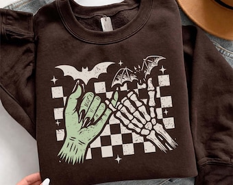 May include: A dark brown crewneck sweatshirt featuring a graphic print of a light green hand and a white skeleton hand performing a pinky promise. The design includes a black and white checkered background, two flying bats, and small stars.