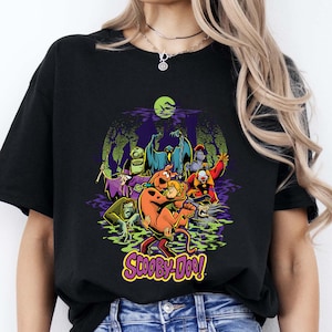 May include: A black t-shirt featuring a colorful graphic print of Scooby-Doo and Shaggy surrounded by various classic villains in a swampy setting under a bright green moon. The text Scooby-Doo is printed in yellow and purple at the bottom.