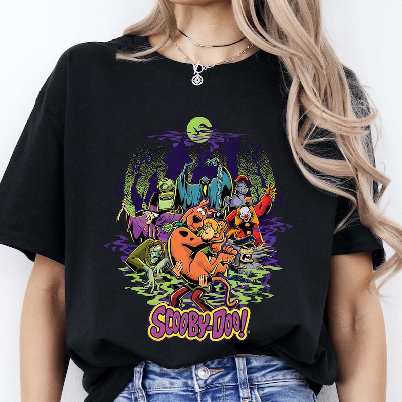 May include: A black t-shirt featuring a colorful graphic print of Scooby-Doo and Shaggy surrounded by various classic villains in a swampy setting under a bright green moon. The text Scooby-Doo is printed in yellow and purple at the bottom.