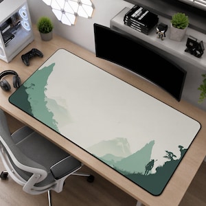 May include: A rectangular desk mat featuring a minimalist landscape illustration in shades of sage green and off-white. The design depicts a silhouette of a person standing on a cliff edge overlooking distant mountains and trees on a light background.