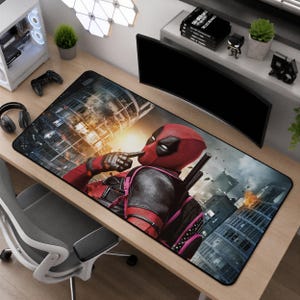 May include: A rectangular desk mat featuring a graphic illustration of the character Deadpool in a red and black suit, set against a backdrop of a burning city skyline with smoke and debris, rendered in shades of red, black, grey, and orange.