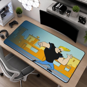 May include: A rectangular desk mat featuring a cartoon illustration of a muscular character with blonde hair wearing a black t-shirt and blue jeans, set against a background of yellow buildings under a blue sky, with a smooth surface finish.
