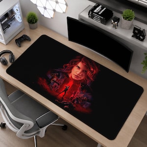 Puede incluir: Una alfombrilla de escritorio rectangular negra que presenta una ilustración gráfica central de una persona con cabello castaño rojizo, rodeada por una escena oscura y atmosférica con iluminación roja vibrante, siluetas de figuras y elementos arquitectónicos.