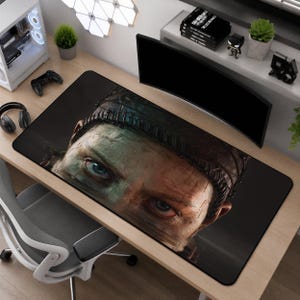 May include: A rectangular desk mat featuring a high-resolution graphic print of a person's face with intense blue eyes and dark, textured headwear. The design covers the entire surface of the mat, which is positioned on a wooden desk.