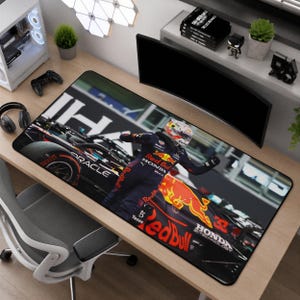 以下が含まれることがあります： ダークブルーのレッドブルレーシングスーツを着たF1ドライバーが、黒と黄色のオラクル・レッドブル・レーシングカーの前に立っている高解像度プリントが施された長方形のデスクマット。レッドブルのロゴとテキストが含まれています。