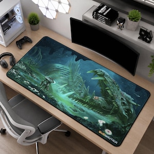 Peut inclure: Un tapis de bureau rectangulaire avec une impression graphique Subnautica. L'illustration montre un grand crâne de créature squelettique dans des tons de vert et de bleu, avec un petit véhicule submersible à proximité. Le mot Subnautica est imprimé en bas.