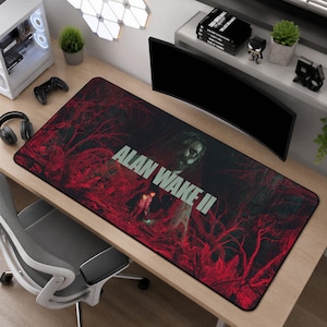Pode incluir: Um tapete de secretária retangular com uma cena de floresta escura em tons de vermelho e o texto ALAN WAKE II impresso em letras brancas grandes. O gráfico inclui uma figura central e ramos de árvore densos e sombrios.
