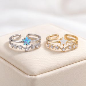 May include: Two open-band rings featuring a star-shaped stone and a row of small round clear stones. One ring is silver-toned with a blue iridescent star, and the other is gold-toned with a white iridescent star, both with a V-shaped design.