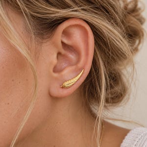 May include: A gold ear climber earring shaped like a delicate feather with detailed veins and a pointed tip. The piece follows the natural curve of the earlobe, featuring a polished metallic finish that catches the light across its textured surface.