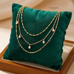 May include: A gold-toned multi-strand necklace featuring delicate twisted chains and four small, clear, pear-shaped drop pendants. The jewelry is displayed on a dark green velvet cushion with the text Corazon JewelCo printed in the upper right corner.