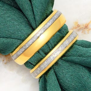 Puede incluir: Dos anillos de tono dorado con una incrustación central de material plateado brillante y texturizado. Las bandas tienen un acabado pulido y reflectante en los bordes exteriores, creando un contraste con el canal central granulado.