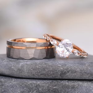 Könnte beinhalten: Ein Set aus zwei Ringen, bestehend aus einem breiten, dunkelgrauen Band mit gehämmertem Muster und einer dünnen, roségoldenen Rille in der Mitte, sowie einem roségoldenen Verlobungsring mit einem großen ovalen Mittelstein und seitlichen Steinen.