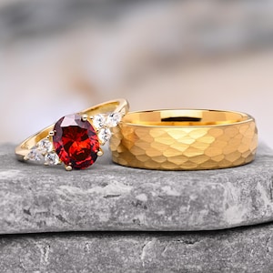Könnte beinhalten: Ein Goldring mit einem großen, oval geschliffenen roten Edelstein, flankiert von kleinen klaren Steinen, kombiniert mit einem breiten Goldring mit gehämmertem Finish. Beide Ringe bestehen aus poliertem Gelbgold mit einer reflektierenden Oberfläche.