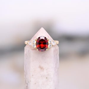 Peut inclure: Une bague en or ornée d'une grande pierre précieuse rouge foncé de taille ovale au centre, flanquée de trois petites pierres blanches claires en forme de poire de chaque côté. La bague est montée sur un anneau en or poli, contrastant les pierres.