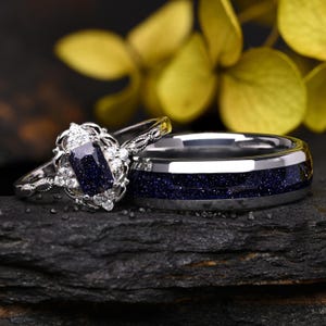 Peut inclure: Un ensemble de bagues argentées comprenant une alliance avec une incrustation de pierre de sable bleu foncé scintillante et une bague de fiançailles avec une pierre centrale rectangulaire bleu foncé entourée de petites pierres claires.