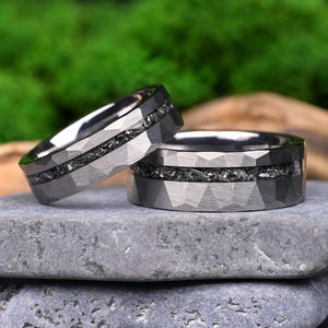 Pode incluir: Dois anéis de metal cinza-escuro com uma superfície exterior geométrica facetada e uma incrustação central de material texturizado, escuro e fragmentado. As bandas têm um interior polido e um perfil largo e plano, exibindo um contraste distinto entre as facetas angulares lisas e a textura granular da incrustação central.