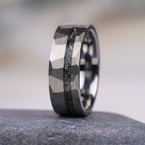 Può includere: Un anello grigio scuro con finitura esterna sfaccettata e martellata, caratterizzato da un inserto centrale in materiale testurizzato e maculato. La fascia ha un profilo largo con superficie interna lucida e un motivo geometrico sull'esterno.