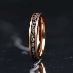 Può includere: Un anello a fascia in oro rosa con un intarsio centrale in meteorite scuro e testurizzato. L'anello presenta una finitura lucida in oro rosa su entrambi i bordi, che incornicia il motivo cristallino ruvido, grigio e nero dell'intarsio.