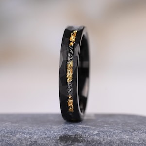 Può includere: Un anello nero con finitura martellata e un inserto centrale caratterizzato da un mix di scaglie color oro e materiale scuro e testurizzato. La fascia presenta un interno lucido e un aspetto robusto su tutta la superficie esterna.