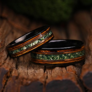 Op de afbeelding: Twee ringen met een zwarte binnenband, een centrale inleg van groene mosagaat en buitenbanden van bruin hout. De ringen hebben een gepolijste afwerking en zijn samen geplaatst, wat de gelaagde combinatie van donker metaal, hout en steen laat zien.
