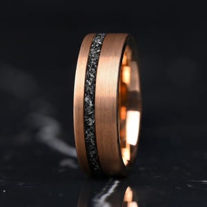 Könnte beinhalten: Ein roségoldfarbener Ring mit gebürsteter Metalloberfläche und einer mittigen Einlage aus einem dunklen, texturierten, kristallinen Material. Das Band hat ein flaches Profil und eine glatte Innenseite, die einen deutlichen Kontrast zwischen der metallischen Oberfläche und der dunklen, gezackten Einlage zeigt, die horizontal um die Mitte des Rings verläuft.