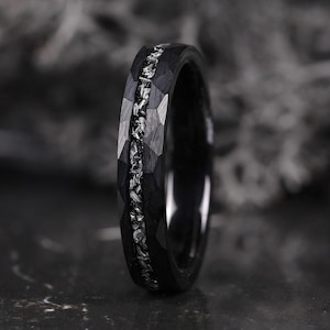 Peut inclure: Une bague noire présentant une texture martelée et facettée sur la surface extérieure, avec une incrustation centrale de fragments métalliques argentés. L'anneau possède une finition sombre et mate avec un canal central réfléchissant.