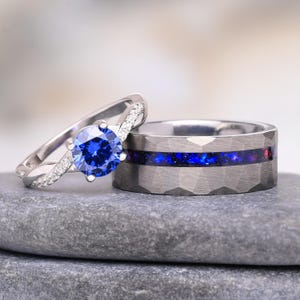 Puede incluir: Un anillo plateado con una piedra central redonda azul y pequeñas piedras claras en una banda trenzada, junto a una alianza ancha plateada y martillada con una incrustación central de material azul y morado iridiscente.