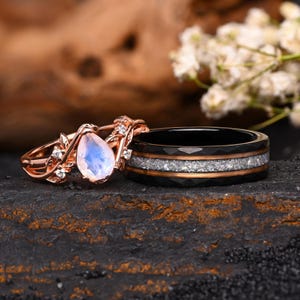 Op de afbeelding: Een roségouden ring met een peervormige maansteen en kleine heldere stenen in een bladvormige band, gecombineerd met een zwarte keramische ring met een centrale inleg van zilverkleurig materiaal en dunne roségouden randen.