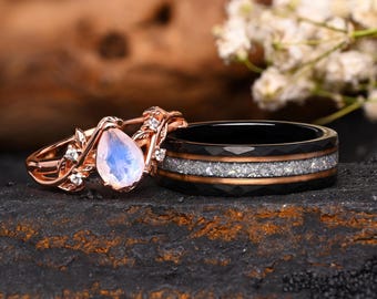 May include: A rose gold ring featuring a pear-shaped moonstone and small clear stones set in a leaf-like band, paired with a black ceramic band featuring a central inlay of crushed silver-toned material and thin rose gold edges.
