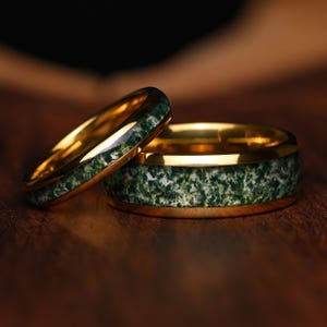 Peut inclure: Deux bagues dorées avec une incrustation centrale en pierre d'agate mousse verte et blanche. Les bagues présentent une finition métallique polie et un design de bande arrondi et lisse, mettant en valeur un motif naturel tacheté.