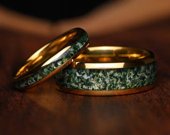 May include: Two gold-toned rings featuring a central inlay of green and white moss agate stone. The rings have a polished metallic finish, with one band being noticeably wider than the other, showcasing the textured, natural green mineral pattern.