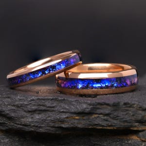 Pode incluir: Dois anéis de ouro rosa com uma incrustação central de material triturado com aparência de galáxia. A incrustação exibe tons vibrantes de azul profundo, roxo e preto, criando um efeito cintilante na superfície de ambos os anéis.