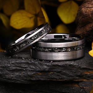 May include: Two silver-toned metal rings featuring a central inlay of dark, textured, fragmented material. The rings have a brushed finish on the outer edges and a smooth, polished interior surface, showcasing a distinct contrast between the metallic bands.