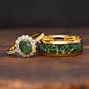Könnte beinhalten: Ein Goldring mit einem oval geschliffenen grünen Moosachat, umgeben von einem Kranz aus kleinen klaren runden Steinen, kombiniert mit einem breiten Goldring, der mit einem texturierten grünen Moosachat rundherum eingelegt ist.
