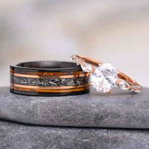 Può includere: Un anello a fascia nero con un inserto centrale in materiale grigio testurizzato fiancheggiato da due sottili strisce color rame, abbinato a un anello di fidanzamento in oro rosa con una grande pietra centrale ovale e pietre laterali marquise.