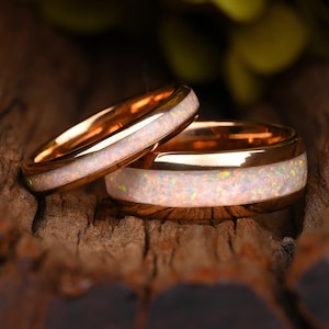 Puede incluir: Dos anillos de oro rosa con una incrustación central de ópalo blanco triturado. La incrustación de ópalo muestra destellos iridiscentes de luz rosa, verde y amarilla. Las bandas de metal tienen un acabado pulido y un perfil redondeado.